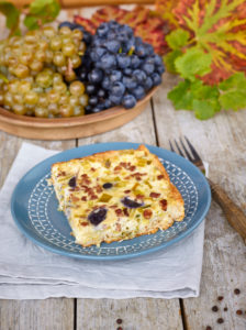 Quiche with grapes and leek, blue country plate on linen cloth, grapes in bowl, fork with wooden handle, table with untreated table top Quiche with grapes and leek, blue country plate on linen cloth, grapes in bowl, fork with wooden handle, table with untreated table top